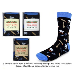 May include: Three boxes of black socks with a blue trim and a variety of kitchen-themed patterns. The boxes have labels with different holiday greetings and names: "Season's Greetings CHARLES", "Merry Christmas BRANDON", and "Happy Holidays ROBERT".