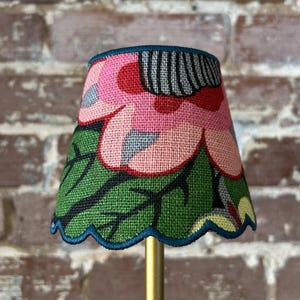 May include: A small, tapered lampshade featuring a vibrant floral pattern with large pink petals, green leaves, and black outlines. The shade has a scalloped bottom edge trimmed in dark blue, set against a textured fabric surface.