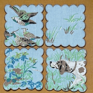 May include: Set of four light blue fabric coasters with scalloped edges. Each coaster features a different nature scene: birds, tall grass, foliage, and a hunting dog. The edges are trimmed with a brown border.