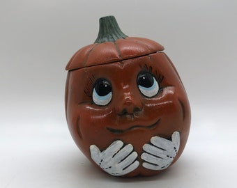 May include: A ceramic pumpkin-shaped cookie jar with a smiling face, large eyes, and white-painted hands. The pumpkin is orange with a green stem and a lid. A whimsical Halloween or fall-themed kitchen decor item.