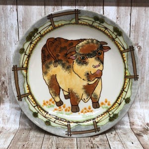 May include: A ceramic plate with a brown and white bull illustration. The plate has a green, brown, and white border with a fence design.