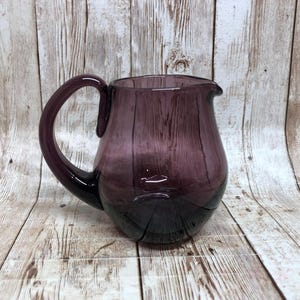 May include: A purple glass pitcher with a rounded body and a large handle. The pitcher has a dark purple base that fades to a lighter purple towards the top. The pitcher is likely used for serving beverages.