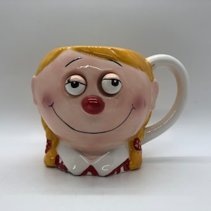 May include: A ceramic mug shaped like a girl's face with a big smile, yellow hair, and large, closed eyes. The mug has a white handle and red and yellow details on the collar.