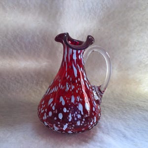 May include: A red glass pitcher with white and blue speckles. The pitcher has a clear glass handle and a ruffled rim. The glass is a deep red color with white and blue spots throughout. The pitcher is a decorative item.
