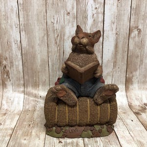May include: A brown rabbit figurine sitting on a hay bale, wearing blue jeans and reading a book. The figurine is titled "Levi".