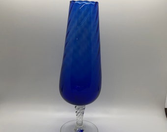 May include: A tall, cobalt blue glass vase with a tapered, ribbed body and a scalloped rim. The vase is supported by a clear, twisted glass stem that connects to a circular, transparent glass base, creating a balanced and elegant silhouette.