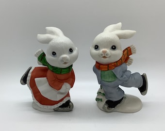May include: Two ceramic bunny figurines. One bunny wears a red dress and green scarf, the other wears a blue outfit and orange scarf. Both are wearing ice skates. The bunnies have white fur and are smiling.