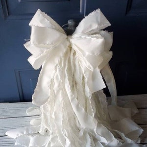 May include: A large, white, ruffled fabric bow with frayed edges. The bow is tied with a loop and is ready to be used as a decorative element.