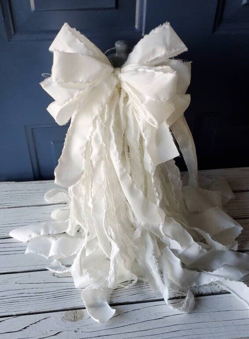 May include: A large, white, ruffled fabric bow with frayed edges. The bow is tied with a loop and is ready to be used as a decorative element.