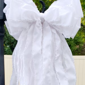 May include: A large, white fabric bow with frayed edges hangs from a black metal hook. The bow is made of layered fabric, creating a textured, voluminous appearance. The background includes greenery and a white fence.