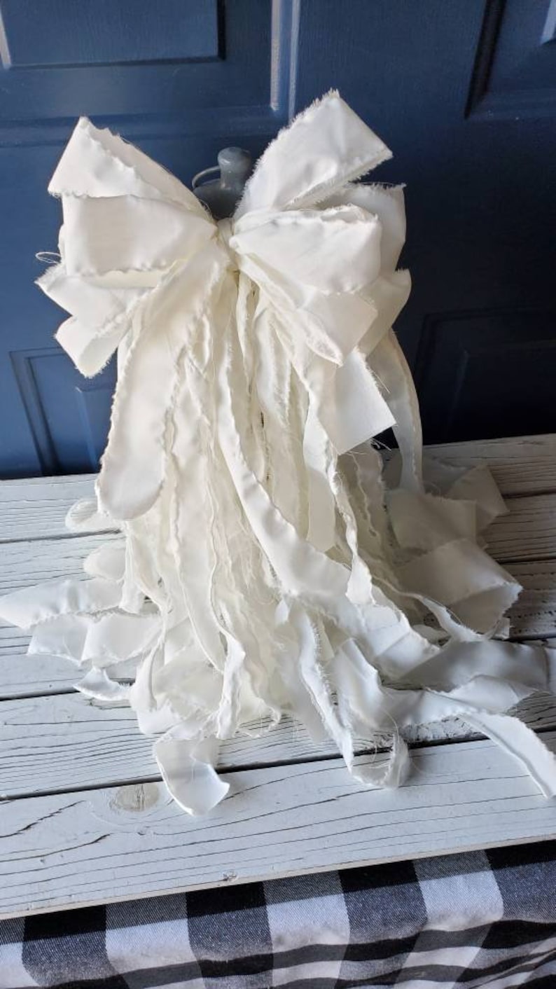 May include: A large white bow with frayed edges and long, flowing tails. The bow is made of fabric and is perfect for adding a touch of elegance to any occasion.