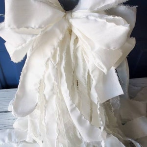 May include: A large white bow made of fabric with frayed edges. The bow is tied with a knot in the center and has long, flowing tails.