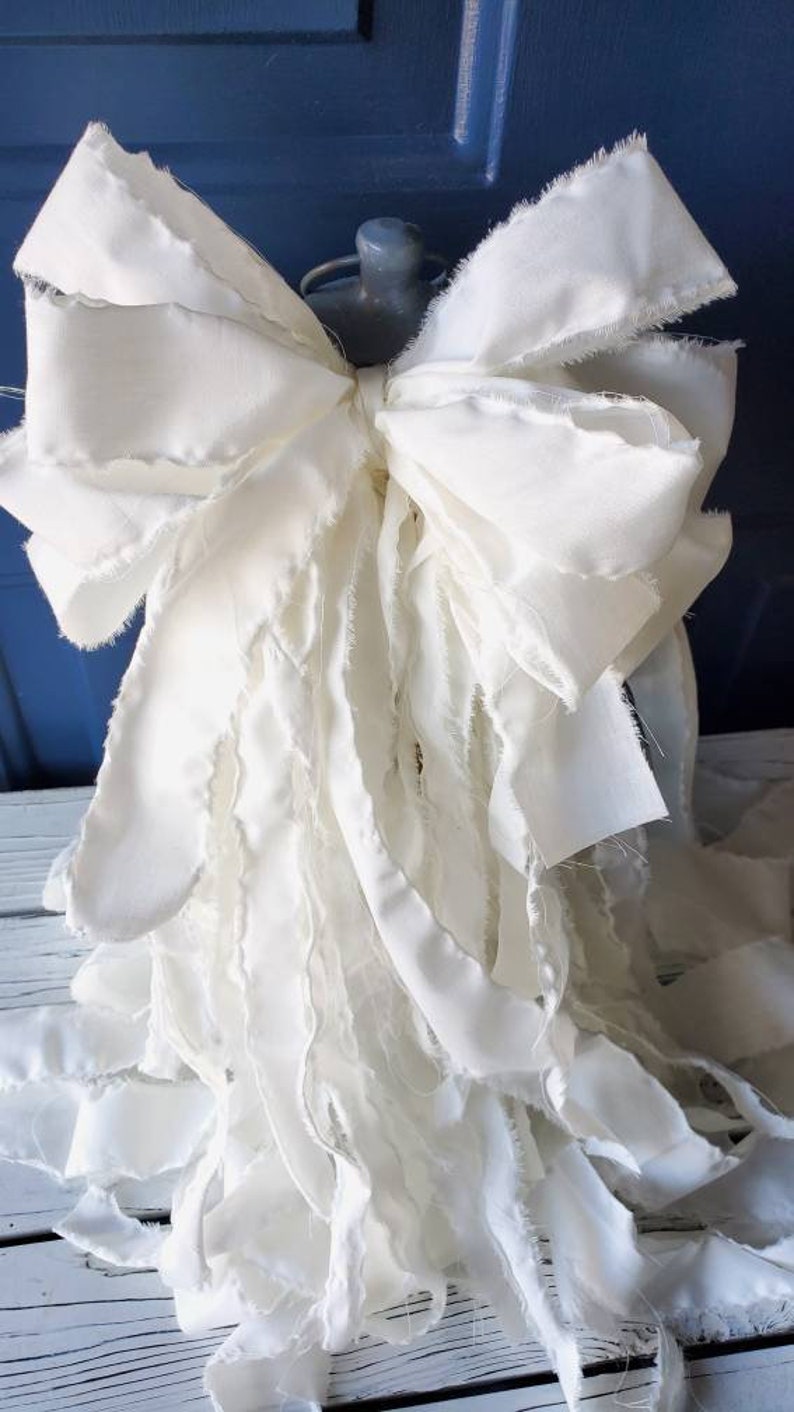 May include: A large white bow made of fabric with frayed edges. The bow is tied with a knot in the center and has long, flowing tails.