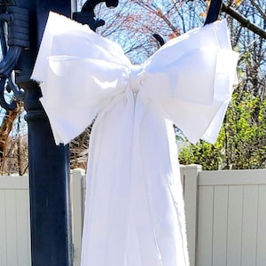 May include: A large, white fabric bow is tied to a black lamppost. The bow has multiple layers of fabric, creating a full, textured appearance. The bow's long, flowing tails cascade downwards. The background includes a white fence and trees.