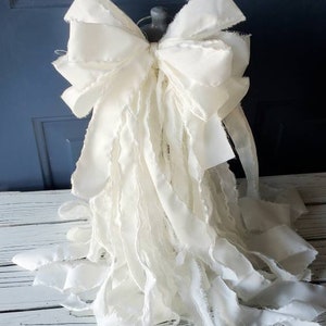 May include: A large, white, frayed fabric bow with a ruffled texture. The bow is tied around a base and is displayed on a white wooden surface.