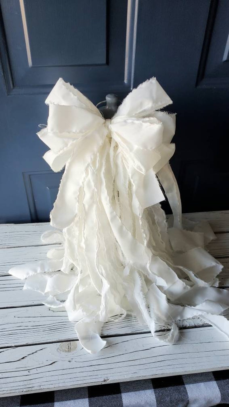 May include: A large, white, frayed fabric bow with a ruffled texture. The bow is tied around a base and is displayed on a white wooden surface.