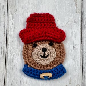 May include: A handmade crochet bear applique. The bear wears a red hat and a blue collar with a wooden button. The bear's face is tan with black eyes and nose. Ideal for crafting or adding to clothing.