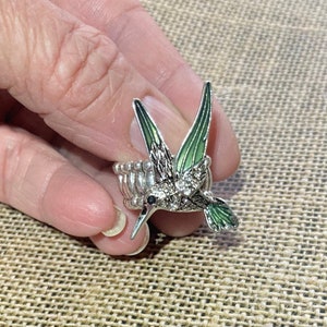 May include: A silver ring with a green and silver hummingbird design. The hummingbird has clear rhinestones on its body.