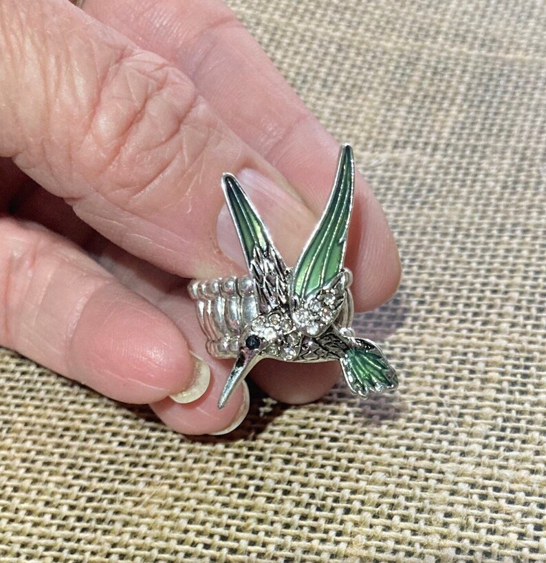 May include: A silver ring with a green and silver hummingbird design. The hummingbird has clear rhinestones on its body.