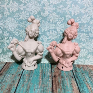 May include: Two ceramic figurines of women with elaborate hairstyles. One figurine is white and the other is pink. Both figurines are wearing dresses with ruffled sleeves and holding a flower in their hands.