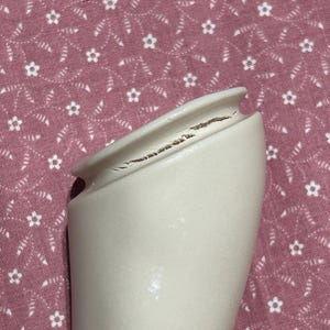 May include: A cream-colored ceramic vase with a cracked rim. The vase has a smooth surface and a flared opening. The background is a pink fabric with a white floral pattern.