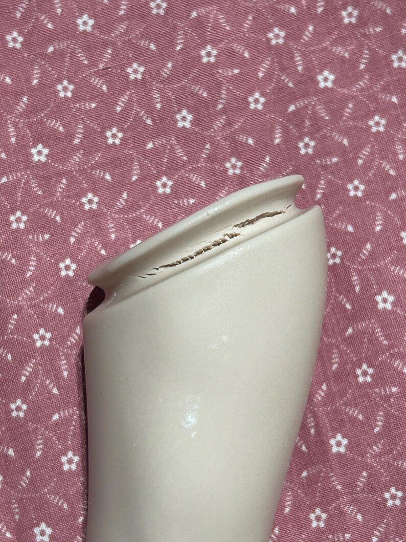 May include: A cream-colored ceramic vase with a cracked rim. The vase has a smooth surface and a flared opening. The background is a pink fabric with a white floral pattern.