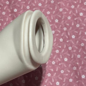 May include: A cylindrical, off-white, smooth-textured object with a rounded, protruding rim at the opening. The object features a hollow center and a uniform, matte finish, showing a simple, functional design with no additional markings or patterns.
