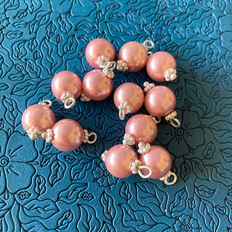 May include: A collection of 10 pink pearl-like ornaments with silver accents. The ornaments have a shiny finish and are arranged on a blue background.