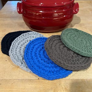 May include: A red ceramic casserole dish with a lid, and a set of five round, knitted coasters in black, gray, blue, dark gray, and green. The coasters are made of thick, textured yarn and are arranged in a semi-circle.