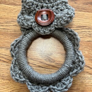 May include: Gray crocheted dishcloth with a wooden button in the center. The dishcloth is shaped like a ring with a scalloped edge.