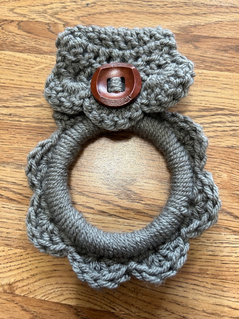May include: Gray crocheted dishcloth with a wooden button in the center. The dishcloth is shaped like a ring with a scalloped edge.