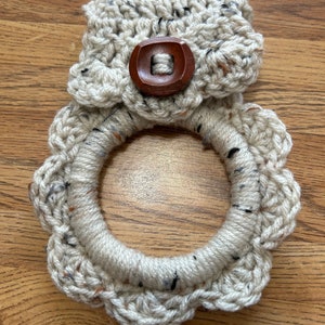 May include: A crocheted dishcloth with a scalloped edge and a brown wooden button. The dishcloth is beige with black and brown flecks.