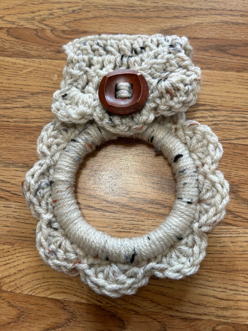 May include: A crocheted dishcloth with a scalloped edge and a brown wooden button. The dishcloth is beige with black and brown flecks.