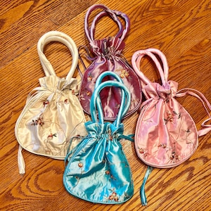 May include: Four drawstring pouches in cream, purple, turquoise, and pink satin fabric. Each pouch features a matching handle, a gathered top, and delicate floral embroidery with small red accents scattered across the surface of the bags.