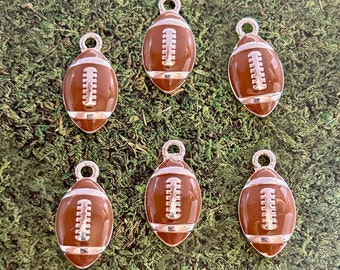 May include: Six brown football charms with silver-colored accents and a loop at the top. The charms are oval-shaped with white stitching details. The background is a textured green surface.