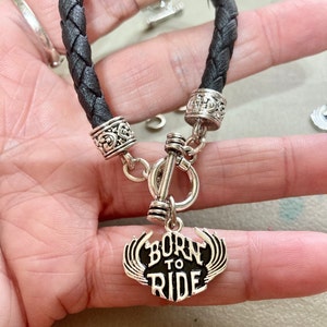 Puede incluir: Pulsera de cuero negro con detalles plateados y un dije que dice "Born to Ride".