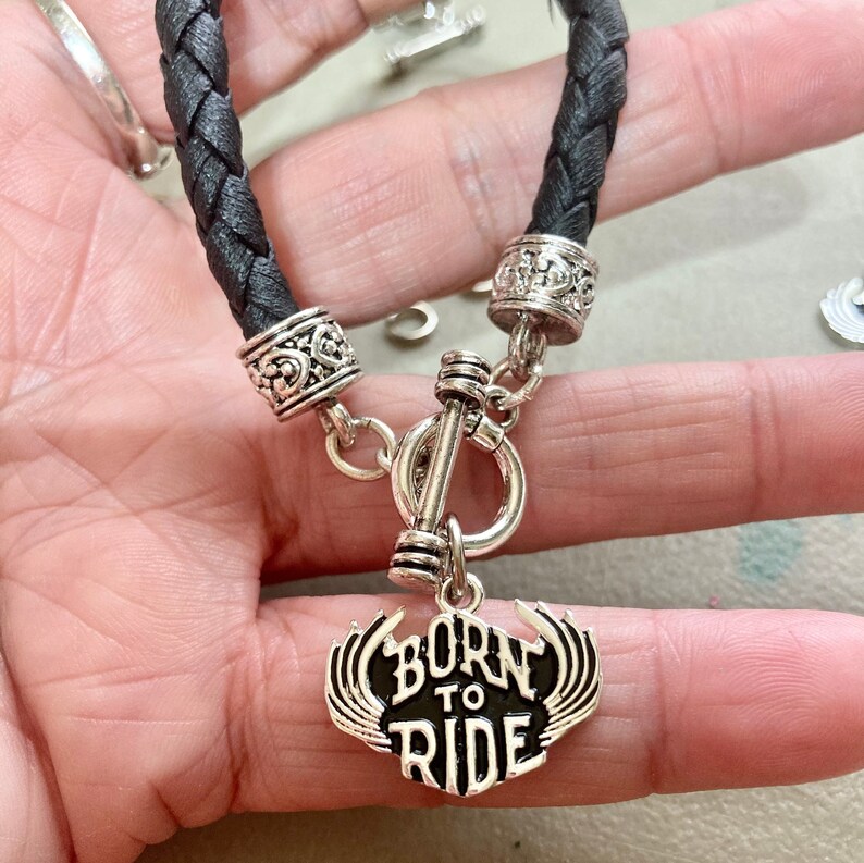Puede incluir: Pulsera de cuero negro con detalles plateados y un dije que dice "Born to Ride".