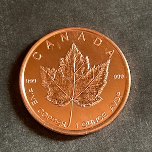 May include: A copper coin with a detailed maple leaf design. The coin is stamped with "CANADA", "FINE COPPER", and "1 OUNCE AVDP". The coin has a diameter of approximately 1.5 inches.