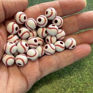 May include: A pile of white baseball-themed beads with red stitching. The beads have a hole through the center for stringing.