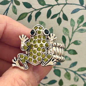 May include: A silver ring with a green jeweled frog design. The frog has black eyes and is surrounded by a silver band.