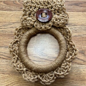 May include: A brown crocheted pot holder with a scalloped edge and a brown button. The pot holder is on a wooden surface.