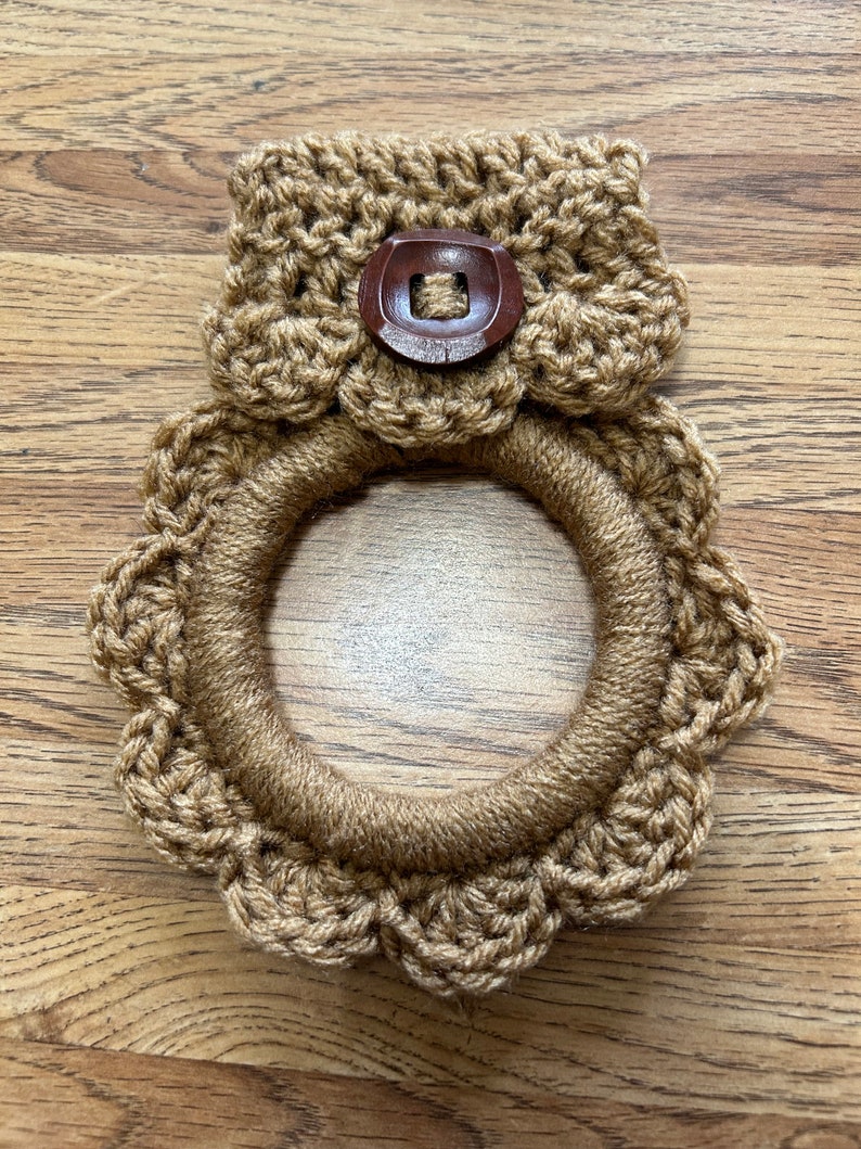 May include: A brown crocheted pot holder with a scalloped edge and a brown button. The pot holder is on a wooden surface.