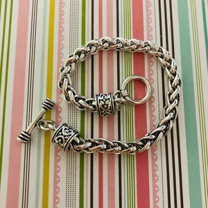 May include: A silver chain bracelet with a toggle clasp and ornate detailing. The bracelet is made of a braided chain and has a decorative ring on one end.