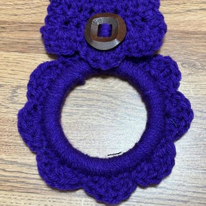 May include: Purple crocheted pot holder with a wooden button. The pot holder is shaped like a flower with a scalloped edge.