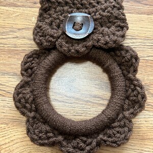 May include: Brown crocheted pot holder with a brown button. The pot holder is shaped like a ring with a scalloped edge.