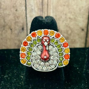 May include: A silver ring with a turkey design. The turkey is made of red, orange, yellow, green, and white rhinestones. The ring is perfect for Thanksgiving or any fall occasion.