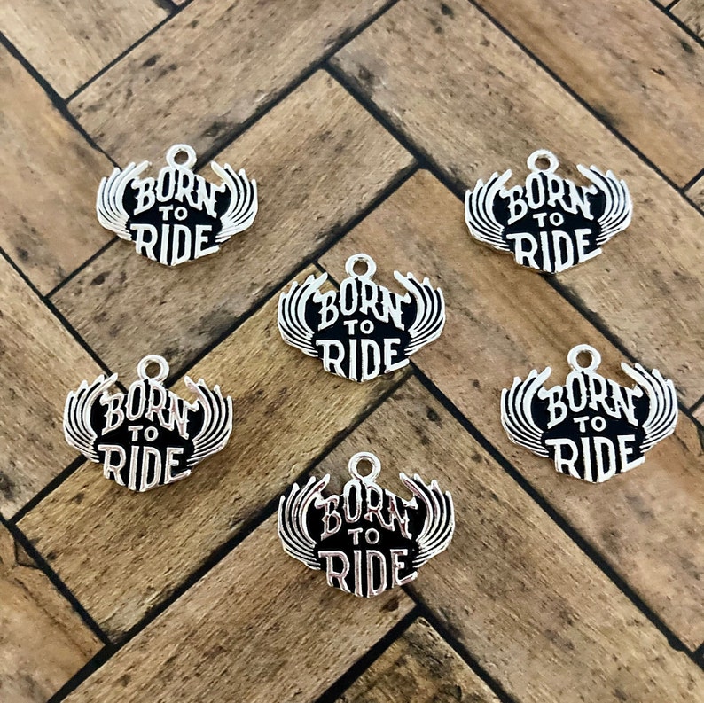 Puede incluir: Seis dijes de plata con letras negras que dicen "Born to Ride" con alas a cada lado.