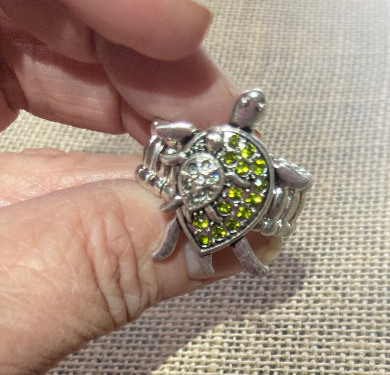 May include: A silver ring with a green and clear crystal turtle charm. The ring is adjustable and has a braided band.