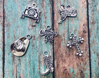 May include: A collection of six silver-toned metal charms featuring a jeep, a horse saddle, a cowboy hat, a cowboy boot, a cross with a rope-like texture, and a script-style charm with the text Country Girl, each with a small jump ring.