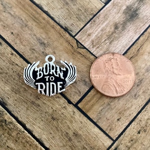 Puede incluir: Encanto de plata con alas negras y las palabras "Born to Ride" en letras blancas. El encanto es aproximadamente del tama&ntilde;o de una moneda de un centavo de d&oacute;lar estadounidense.